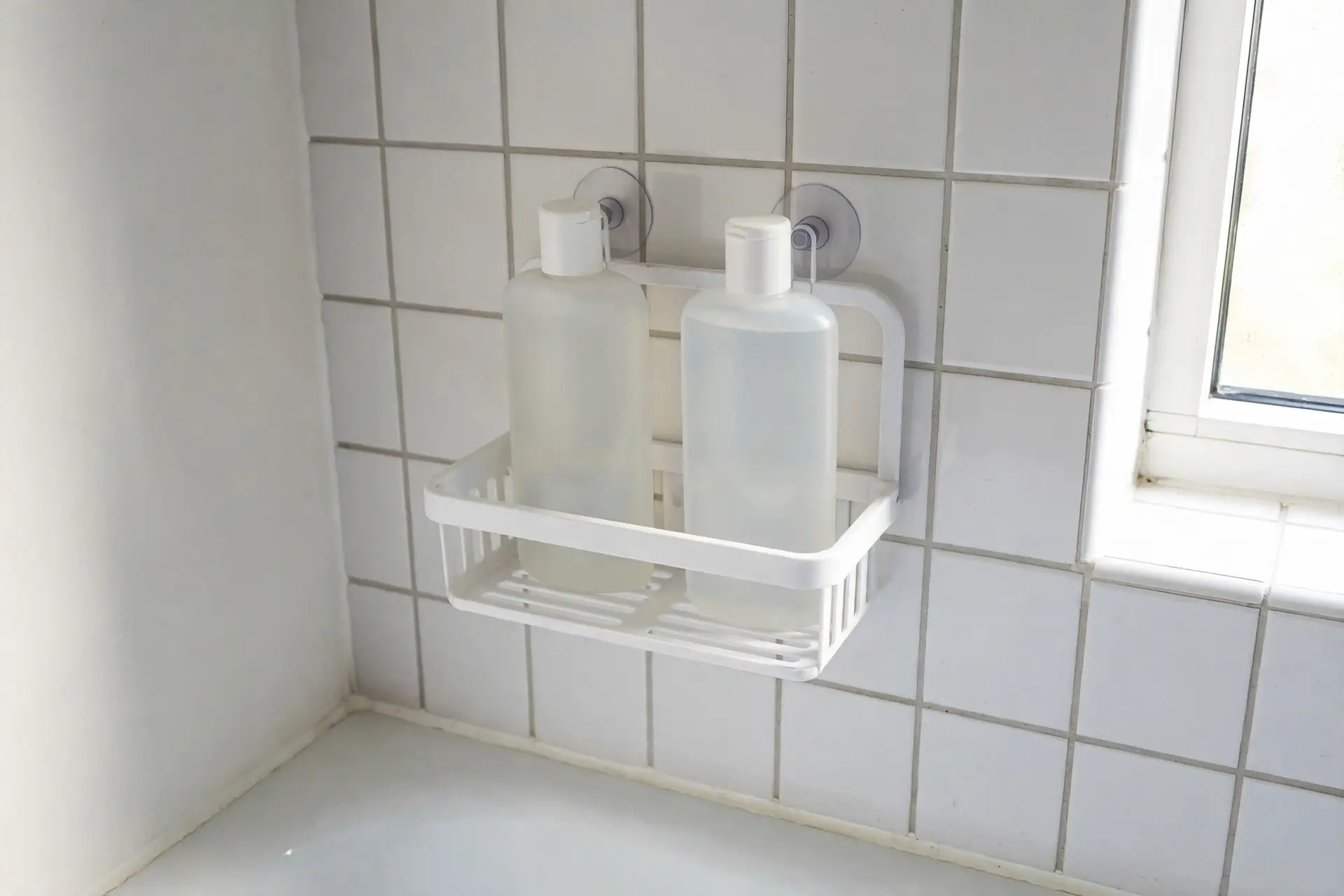White suction-cup shower caddy mounted on bathroom tile holding shampoo and conditioner bottles, with slatted drainage bottom
